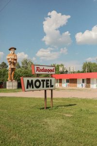 Sunny day at Badlands National Park showing a red roadside motel, a large sign, and a wooden cowboy statue.