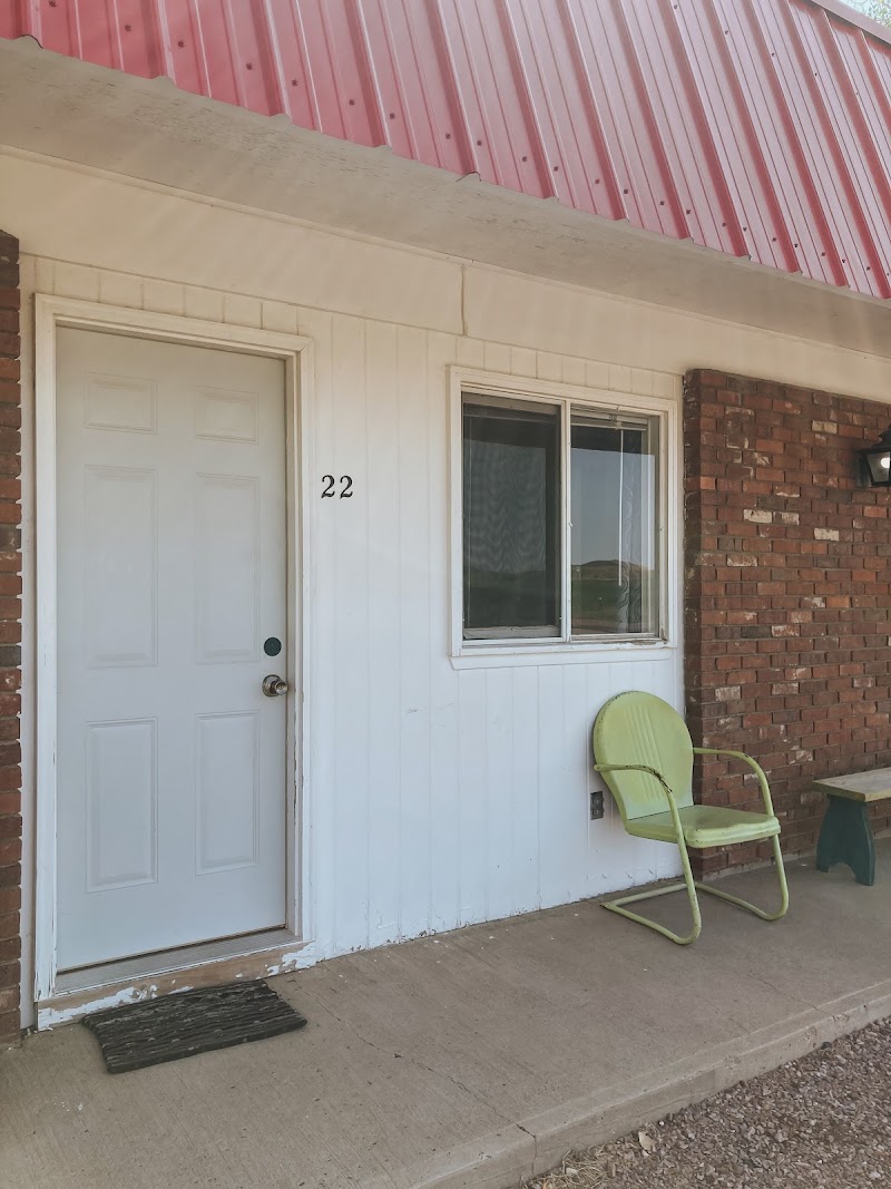 Lodging unit exterior with white siding, door 22, window, lime-green chair on a concrete porch, pink metal roof, Badlands National Park.