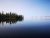 Calm Lewis Lake in Yellowstone National Park, with glassy water reflecting a tree-lined shore and blue sky.