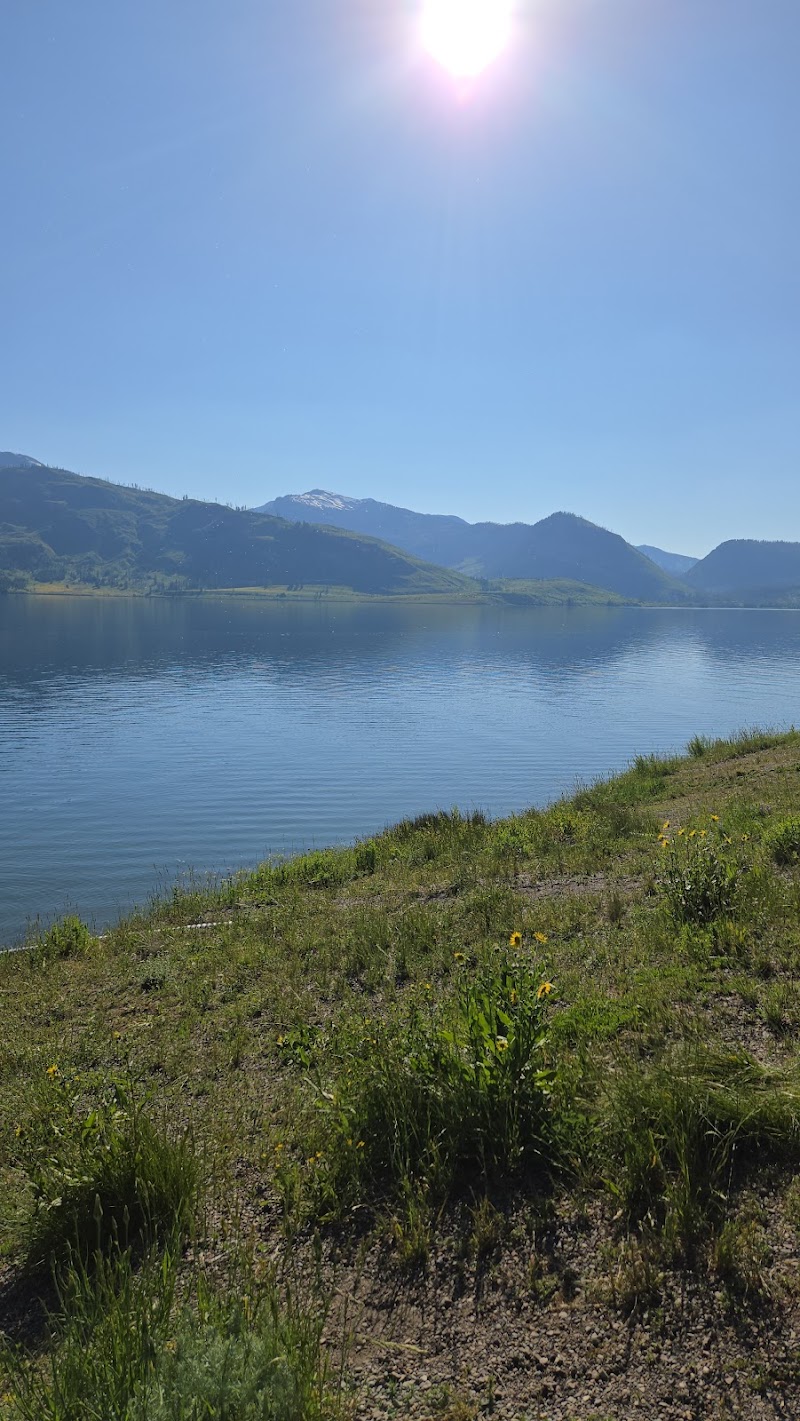 Calm Lewis Lake mirrors the blue sky with grassy shoreline and distant snow-dusted mountains in Yellowstone National Park.