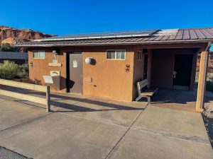 Brown campground facility with two doors and a bench, solar panels on the roof, red rock cliffs at Arches National Park.