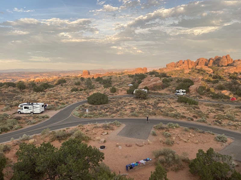 Arches National Park campground scene with a winding road, several RVs, a lone walker, and distant red rock formations.