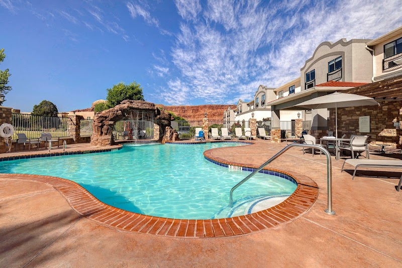 Sunny pool area with brick coping, lounge chairs, and a rock arch in the background at Arches National Park resort.