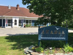 Exterior view of a lodge at Acadia Village Resort in Acadia National Park, with American flag, blue siding, and green trees in bright summer light.