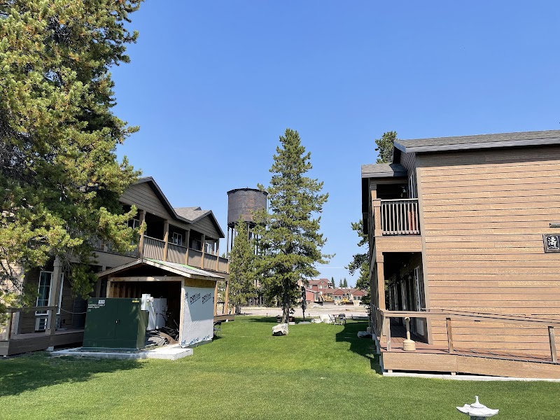 Wood lodge buildings with balconies flank a green lawn, tall pines, and a water tower in Yellowstone National Park.