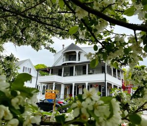 White Victorian inn with wraparound porch and red stairs, framed by blooming white blossoms in Acadia National Park.
