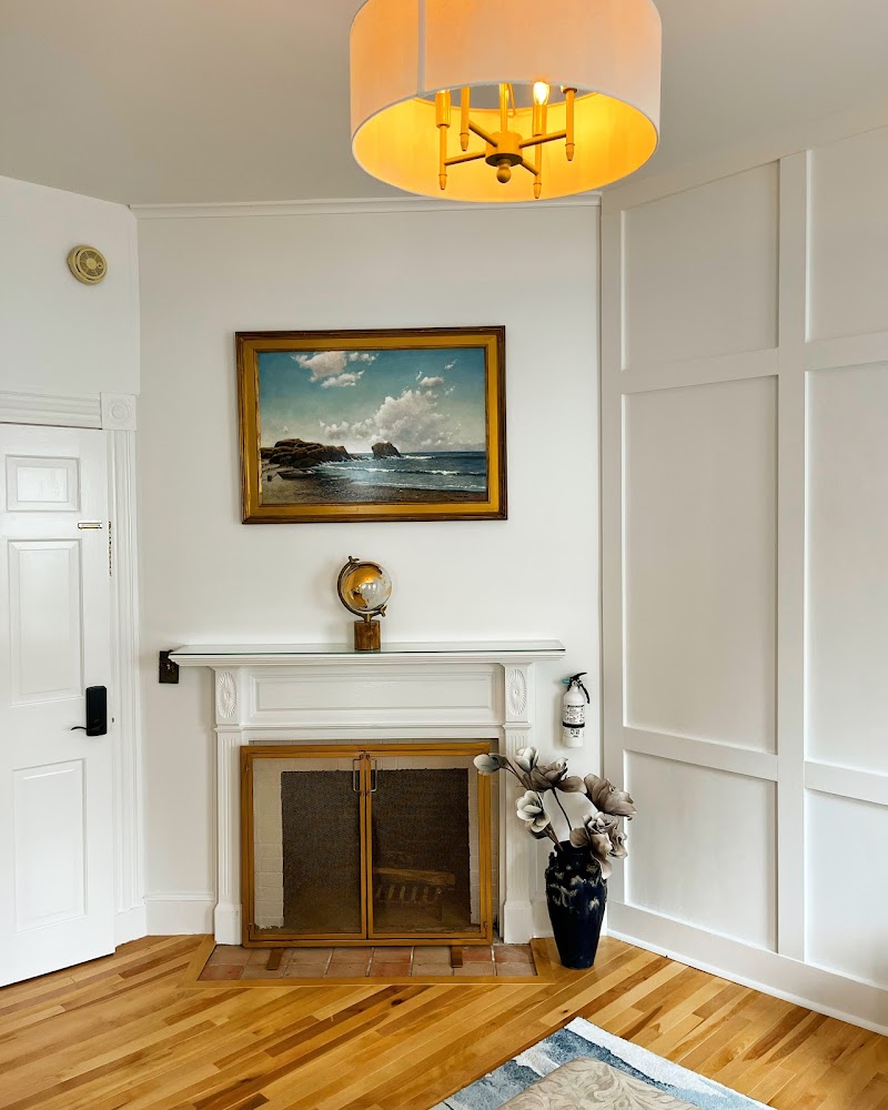 Cozy white living area with a mantel fireplace, framed ocean scene, brass globe clock, tall vase of flowers, and warm ceiling light in Acadia National Park.