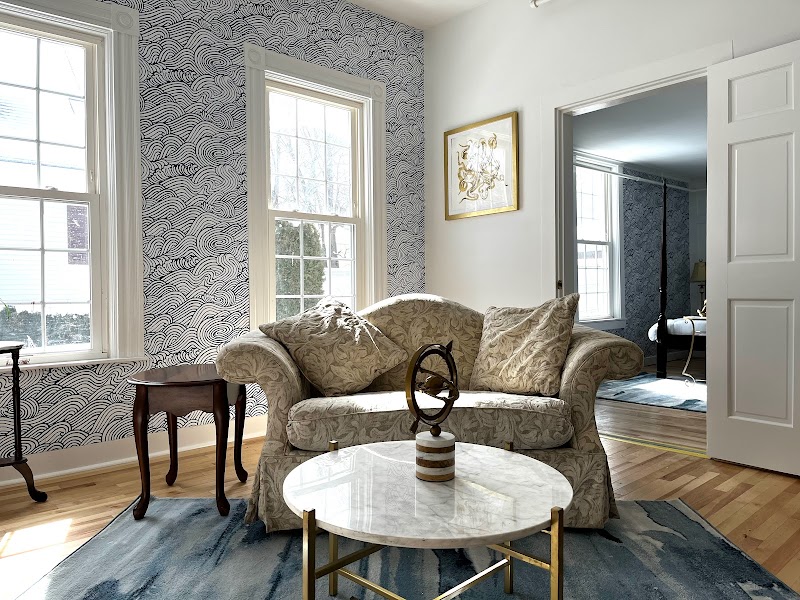 Cozy living room with blue patterned wallpaper, beige sofa with pillows, marble coffee table, two windows, and hardwood floors in Acadia National Park lodging.