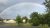 Vivid rainbow arches over a campground in Acadia National Park, with green trees, a yellow building, and a gravel road.