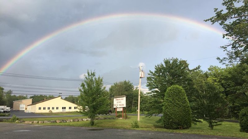 Vivid rainbow arches over a campground in Acadia National Park, with green trees, a yellow building, and a gravel road.