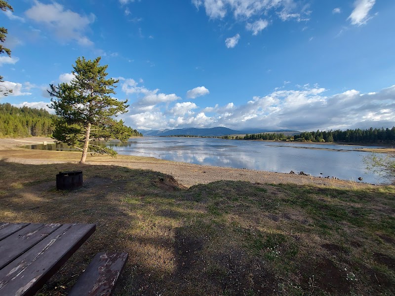 Yellowstone National Park: wooden picnic table beside a lone tree, calm lake, and distant mountains under a blue sky.