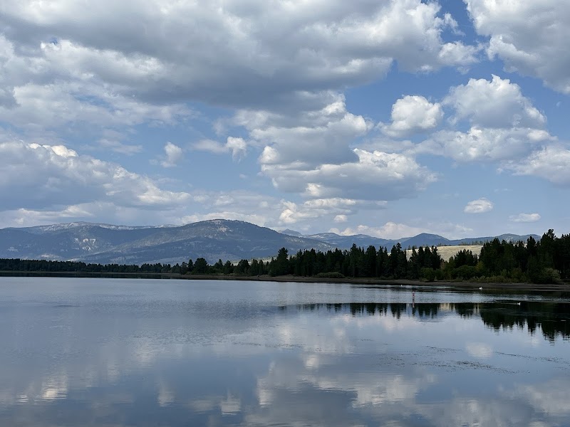 Calm lake in Yellowstone National Park reflecting a tree-lined shore, distant mountains, and a partly cloudy sky.