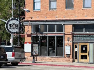 Brick storefronts line a city street with large windows, a round hanging sign, an OPEN sign, and parked SUVs in Arches National Park.