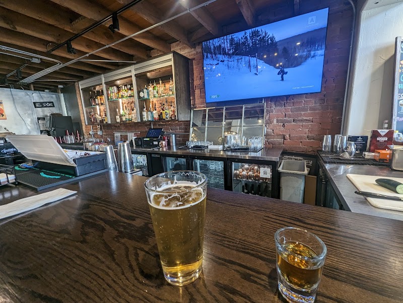 Rustic bar interior at Arches National Park with a wooden counter, a glass of beer and a shot, and shelves of bottles behind a brick wall.