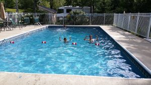 Pool at a campground in Acadia National Park, two swimmers raising arms, red-white buoys, white fence, and trees.