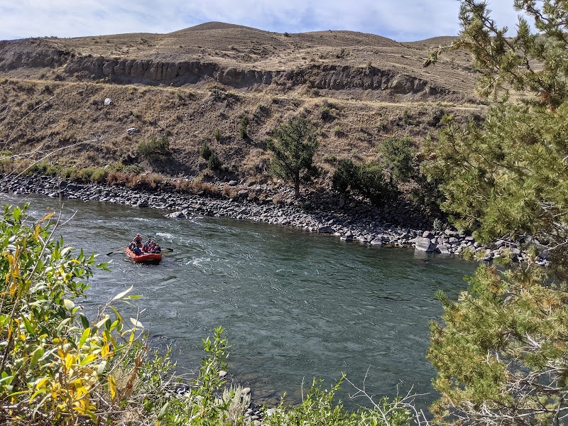 A red inflatable raft carries passengers down a wide river, rocky banks and arid hills in Yellowstone National Park.