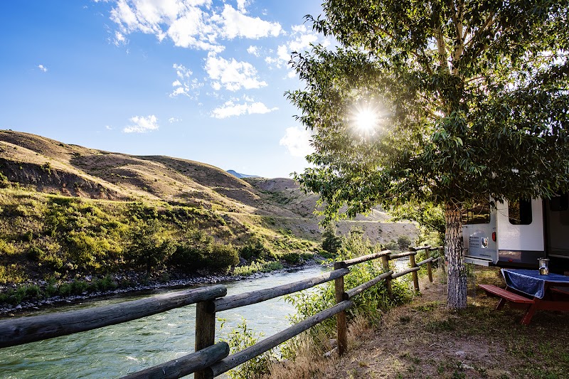 Riverside campground in Yellowstone National Park with wooden fence, RV under a tree, and sunlit hills.