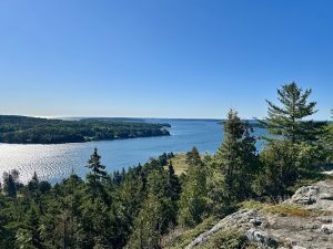 Overlook from Flying Mountain Trailhead in Acadia National Park: rocky ledge, evergreen trees, and a sunlit blue bay.