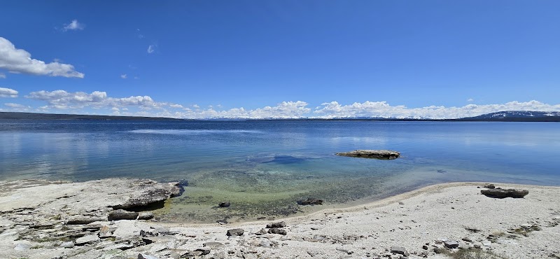 Clear blue water at Yellowstone National Park's Yellowstone Lake from West Thumb Overlook, rocky sandy shore, exposed rocks, distant mountains under a bright sky.