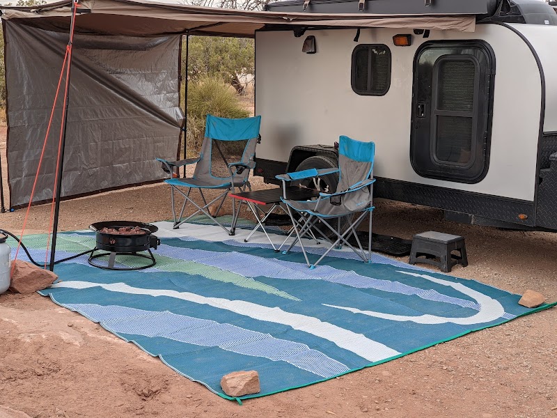 Arches National Park campsite with a white trailer, blue camping chairs, a striped rug, and a small fire pit on desert soil.