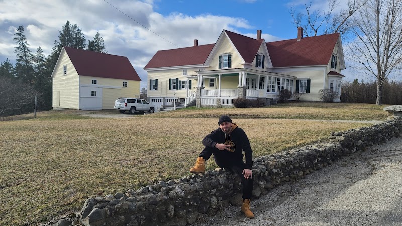 Person kneels on a low stone wall in front of a yellow two-story house with a red roof and white porch at Hancock, Acadia National Park.