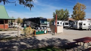 Arches National Park campground scene with a large black motorhome, multiple white RVs, a green-roofed building, gravel lot, and a red picnic table nearby.