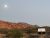 Crescent moon above sunset-red rock cliffs, scrubby desert, and a white tent shelter in Arches National Park.