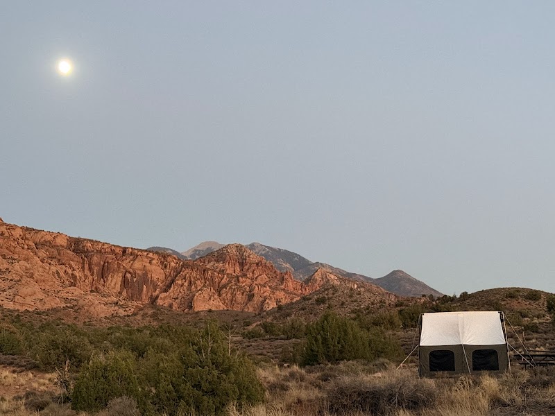 Crescent moon above sunset-red rock cliffs, scrubby desert, and a white tent shelter in Arches National Park.
