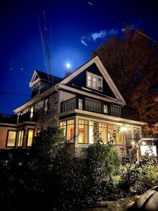 Exterior of a warmly lit lodge in Acadia National Park at night, with a wraparound porch and autumn foliage visible.