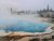 Blue geothermal pool with rising steam along a boardwalk edge, with pines in the distance, Yellowstone National Park.
