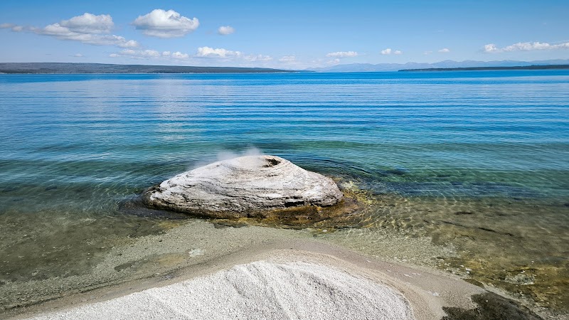 Calm Yellowstone National Park lake with a steaming, milky-white rock on a shallow shore and clear blue water.
