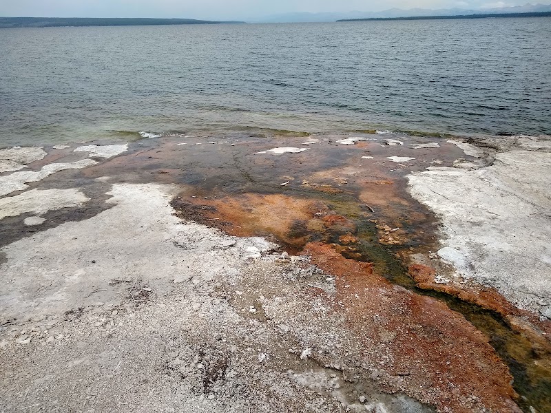 Rocky shoreline with orange, brown, and white mineral deposits fading into a calm lake in Yellowstone National Park.