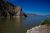 Calm lake with jagged rocky cliffs on the left, blue sky, distant mountains, and a rocky shore in Yellowstone National Park.
