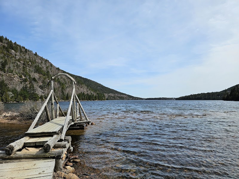 Jordan Pond Path Trailhead at Acadia National Park, a wooden boardwalk extending toward Jordan Pond beside rocky shores under a blue sky.
