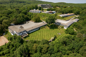 Aerial view of a long, L-shaped lodge complex with a manicured lawn, surrounded by dense pines in Acadia National Park.