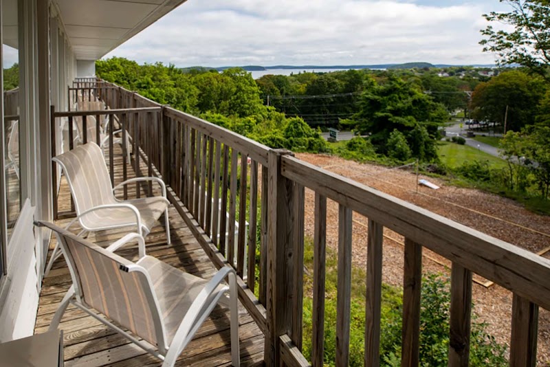 Balcony with white chairs and a weathered railing overlooks green trees, a distant road, and water in Acadia National Park.
