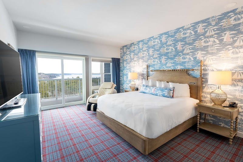 Bright hotel room in Acadia National Park with a wooden bed, sail wallpaper, plaid carpet, and balcony view.