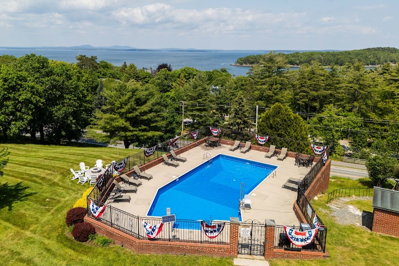 Blue rectangular pool with brick surround, lounge chairs and bunting, surrounded by lush trees with the ocean beyond in Acadia National Park.
