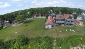 Aerial view of red-roofed cottages and a multi-story lodge on a grassy bluff in Acadia National Park, with trees, lawn chairs, and coastal shoreline nearby.