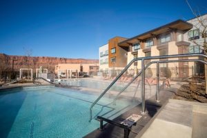 Outdoor hotel pool with metal handrail, lounge chairs, and modern buildings against red rock cliffs in Arches National Park.