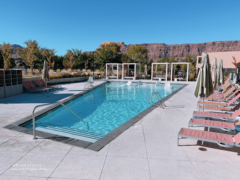 Sunlit pool deck with a rectangular blue pool, pink lounge chairs and umbrellas, with Arches National Park cliffs beyond.