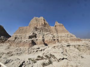 Towering beige badlands rock formation with layered strata beneath a blue sky, railing and hikers on the left at Badlands National Park.