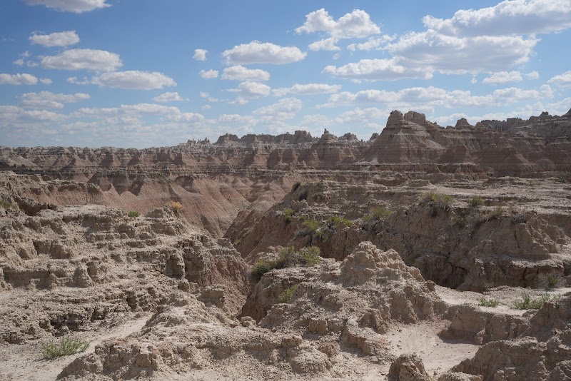 Rugged Badlands National Park terrain with layered eroded buttes, gullies, and sparse vegetation under a blue sky.