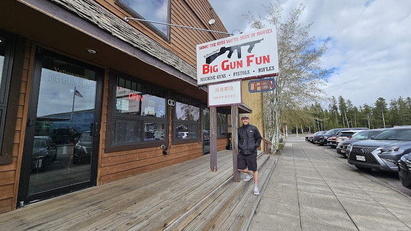 A man in a black jacket and shorts stands on a wooden deck outside a rustic lodge with a large sign in Yellowstone National Park.