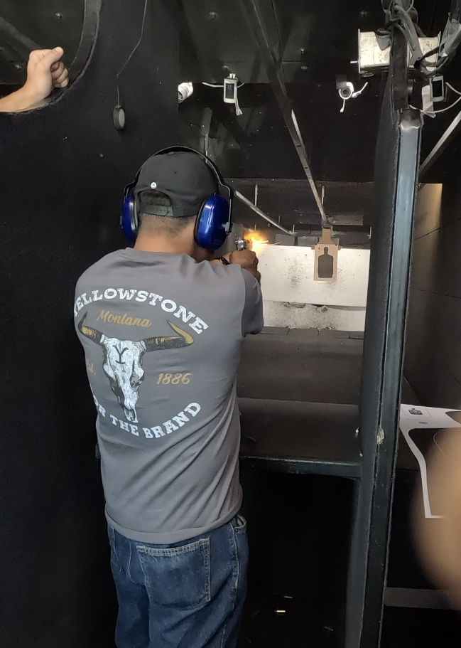 Person in a gray Yellowstone National Park shirt with blue ear protection fires a handgun at an indoor range; silhouette target on a dark wall.