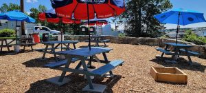 Picnic tables with red and blue umbrellas dot an outdoor dining area in Acadia National Park, gravel ground and bright sky.