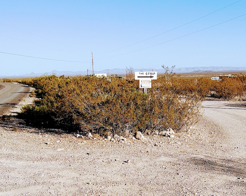Desert road scene with a sign pointing to The Strip amid scrubby bushes in Big Bend National Park at a remote campground area.