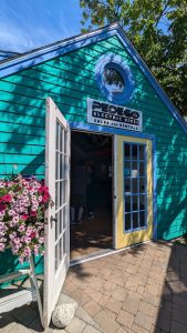Teal gift shop with a round window, yellow-trimmed doors, pink flowers, and visitors inside at Acadia National Park.