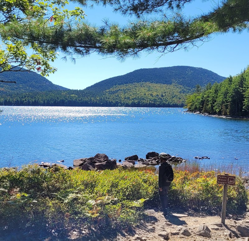 Hiker with backpack beside a blue lake and rocky shore in Acadia National Park, with pine trees and distant hills.