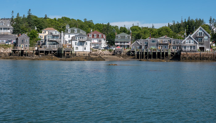 Stonington waterfront houses line the rocky shore along Acadia National Park, with calm blue waters under a bright summer sky.